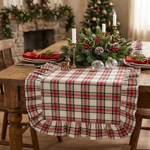 Pode incluir: Um caminho de mesa retangular com padr&atilde;o xadrez vermelho, verde e branco com bordas franzidas. O caminho de tecido est&aacute; sobre uma mesa de madeira, servindo de base para um centro de mesa de ramos verdes, bagas vermelhas e velas.