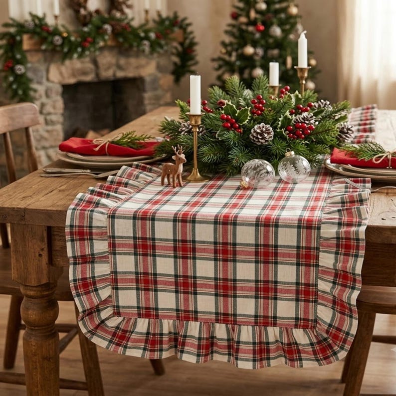 Pode incluir: Um caminho de mesa retangular com padr&atilde;o xadrez vermelho, verde e branco com bordas franzidas. O caminho de tecido est&aacute; sobre uma mesa de madeira, servindo de base para um centro de mesa de ramos verdes, bagas vermelhas e velas.
