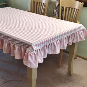 May include: A pink and white gingham tablecloth with a ruffled edge. The tablecloth is draped over a wooden table, creating a classic and charming look. The fabric has a delicate, feminine aesthetic.