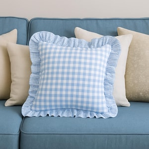 Può includere: Un cuscino a quadretti blu e bianco con bordo a balze è posto su un divano blu, circondato da altri cuscini decorativi dai toni neutri. Il cuscino centrale presenta un classico motivo a quadretti.
