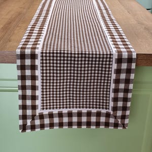 May include: Brown and white checkered table runner with a white border. The runner is made of fabric and has a checkered pattern.