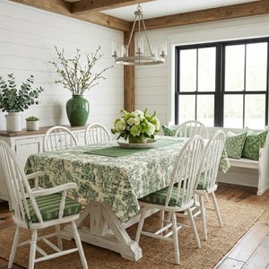 May include: A dining room set featuring a rectangular wooden table covered with a green and white floral toile tablecloth, surrounded by six white spindle-back chairs with green striped cushions and a white wooden bench with green patterned pillows.