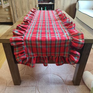 Pode incluir: Um caminho de mesa retangular com um padr&atilde;o xadrez em vermelho, verde e branco. O caminho tem folhos de tecido franzido ao longo de ambas as bordas longas, criando uma borda decorativa. O tecido exibe um design cl&aacute;ssico de tartan.