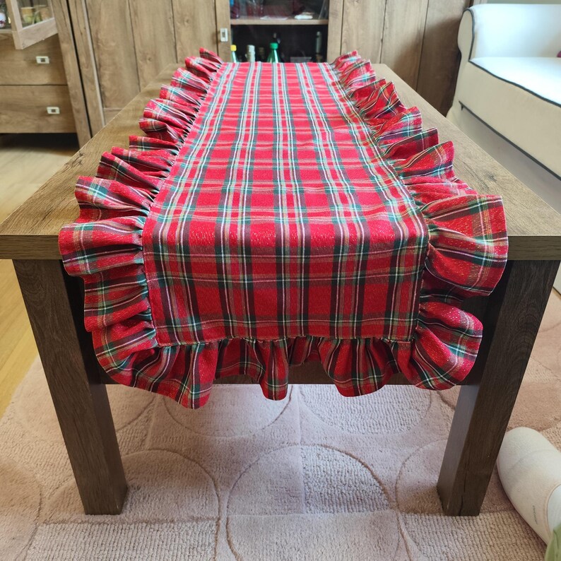 Pode incluir: Um caminho de mesa retangular com um padr&atilde;o xadrez em vermelho, verde e branco. O caminho tem folhos de tecido franzido ao longo de ambas as bordas longas, criando uma borda decorativa. O tecido exibe um design cl&aacute;ssico de tartan.