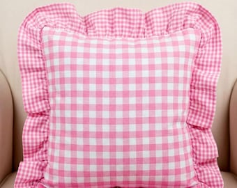 Könnte beinhalten: Ein quadratisches Kissen mit einem rosa-weißen Vichy-Karo-Muster. Das Kissen ist mit einem breiten, gerafften Rüschenrand aus demselben rosa-weiß karierten Stoff versehen, der eine weiche, strukturierte Kante um das Kissen herum erzeugt.