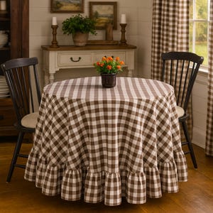 May include: A round table covered with a brown and white checkered tablecloth featuring a ruffled hem. The tablecloth drapes over the edges of the table, which is positioned between two black wooden chairs with vertical slats and beige cushions.