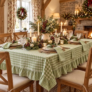 May include: A dining table covered with a green and white gingham tablecloth featuring a ruffled edge. The table is set with white plates, patterned napkins, clear glassware, and a centerpiece of pine branches, red berries, and candles.