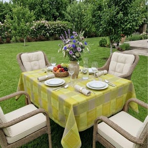 Puede incluir: Una mesa de comedor cuadrada para exteriores con un mantel de cuadros amarillos y grises, dispuesta con cuatro platos blancos, servilletas enrolladas, cristalería y un jarrón central de cerámica con un ramo de flores silvestres y frutas.