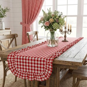 Pode incluir: Uma mesa de jantar rústica é decorada com um caminho de mesa xadrez vermelho e branco com uma borda com babados. Cortinas combinando emolduram uma janela, e um vaso de flores e um castiçal completam a cena. A mesa é de madeira.