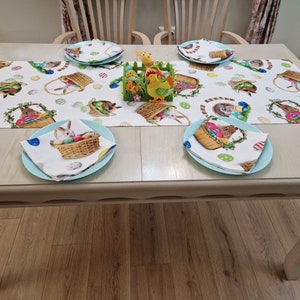 Op de afbeelding: Een lichtblauwe tafelrunner met een witte achtergrond en een herhalend patroon van konijnen die manden met kleurrijke paaseieren vasthouden. De runner ligt op een lichte houten tafel met vier couverts, elk met een lichtblauw bord en een witte servet met een konijn en mand met eieren ontwerp.