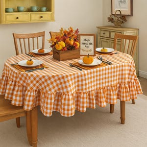 Peut inclure: Une table ovale dressée pour une célébration de la récolte d'automne. La table est recouverte d'une nappe à carreaux orange et blanc avec un bord à volants. Les centres de table comprennent des citrouilles, du feuillage d'automne et une pancarte encadrée.