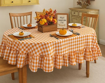 May include: An oval dining table set for a fall harvest celebration. The table is covered with an orange and white checkered tablecloth with a ruffled edge. Centerpieces include pumpkins, fall foliage, and a framed sign that reads "Happy Thanksgiving".