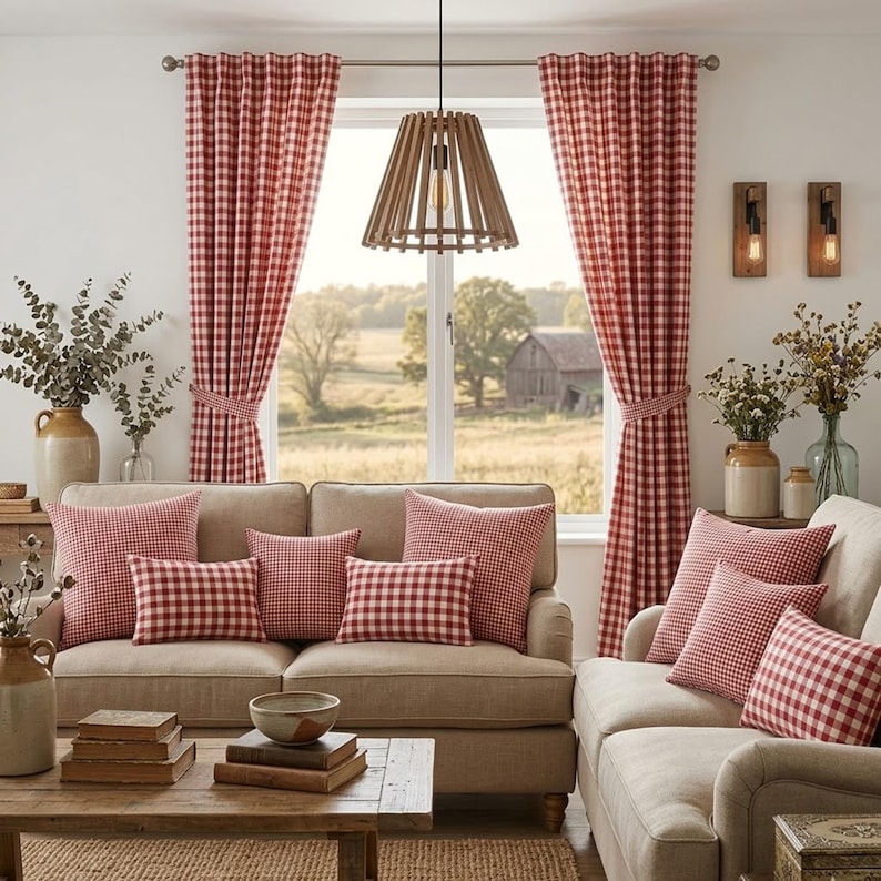 May include: A cozy living room scene featuring two beige sofas adorned with red and white checkered pillows. Red and white checkered curtains frame a window overlooking a rural landscape. A wooden pendant light hangs above a wooden coffee table.