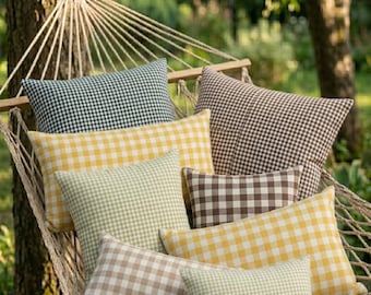 Op de afbeelding: Een verzameling kussens met ruitpatroon in verschillende kleuren, waaronder geel, groen, bruin en zwart, is gerangschikt in een hangmat. De kussens bieden een gezellige en uitnodigende uitstraling, perfect voor ontspanning in de buitenlucht. De hangmat hangt tussen bomen.