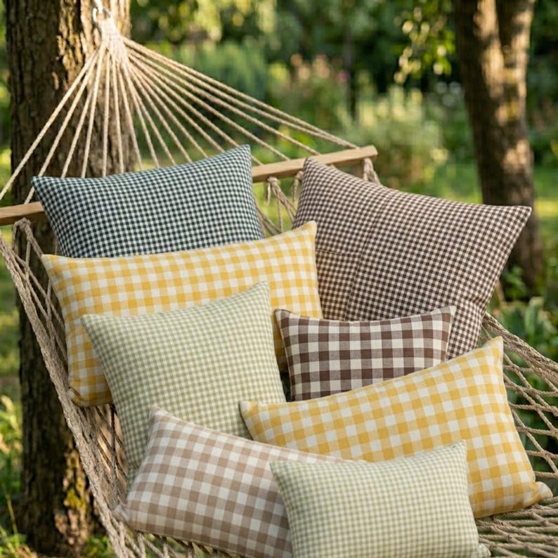 May include: A collection of gingham-patterned pillows in various colors, including yellow, green, brown, and black, are arranged in a hammock. The pillows offer a cozy and inviting aesthetic, perfect for outdoor relaxation. The hammock is suspended between trees.