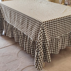 May include: A rectangular tablecloth featuring a brown and white gingham check pattern. The fabric covers the top of a table and includes a gathered, ruffled skirt that hangs down from the edges, creating a layered and textured appearance.
