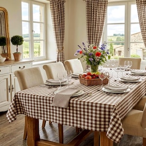 May include: A dining table set with a brown and white checkered tablecloth and matching curtains. The table features beige upholstered chairs, a woven basket filled with colorful wildflowers and fruit, and place settings with white plates and napkins.