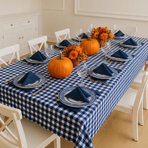May include: A dining table set with a blue and white chequered tablecloth, white plates with blue patterns, navy blue folded napkins, clear wine glasses, and two orange pumpkins surrounded by orange flowers as centrepieces.