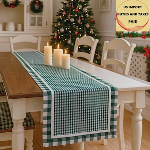 May include: A green and white checkered table runner with a white border, placed on a wooden table. Three lit candles are on the runner. The table is set for a holiday meal, with a Christmas tree in the background.