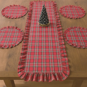 Pode incluir: Um caminho de mesa retangular e quatro jogos americanos circulares com um padr&atilde;o xadrez vermelho, verde e branco com bordas franzidas. Um pequeno ornamento decorativo de &aacute;rvore em forma de cone verde escuro est&aacute; no centro do caminho.