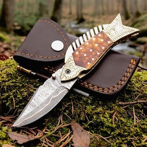 May include: A folding pocket knife shaped like a fish with a patterned metal blade and a brown handle featuring a diamond grid design. The knife is resting on a dark brown leather sheath with a silver snap button closure and visible stitching.