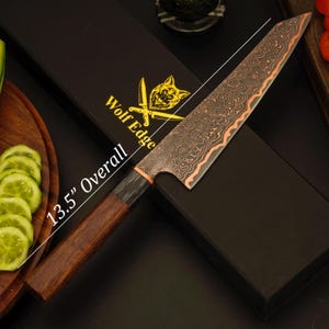 May include: A chef's knife, overall length 34.3 cm, with a patterned blade and wooden handle. The blade has a copper-colored edge and a decorative pattern. The knife is displayed with a black box that has the text "Wolf Edge".