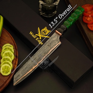 Puede incluir: Un cuchillo de chef de 34,3 cm de largo con una hoja estampada y un mango verde. El cuchillo tiene una virola de color cobre y se muestra con una caja negra con el texto "Wolf Edge". Pepinos y tomates en rodajas en el fondo.