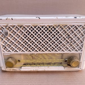 May include: A vintage white Philips radio with a rectangular shape and a prominent diamond-patterned speaker grille. The front panel features a horizontal tuning dial with printed frequency text and two circular control knobs on the lower left and right sides. The white casing shows signs of paint wear and aging.