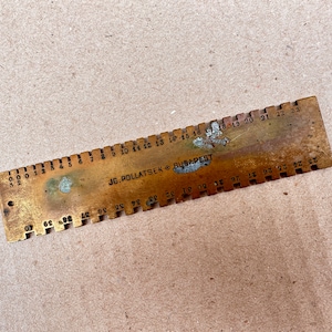 Può includere: Un righello in ottone con una scala graduata da 0 a 23. Il righello è contrassegnato dal testo "JG. POLLATSEK BUDAPEST".