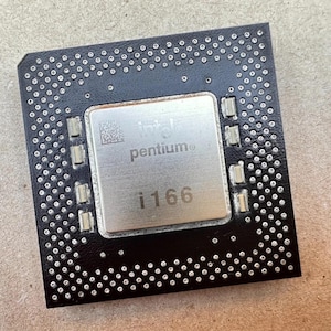 Pode incluir: Um processador de computador preto quadrado com um dissipador de calor central de metal prateado. O texto intel pentium e i166 está impresso na superfície, juntamente com um pequeno código QR quadrado. As bordas apresentam uma grade de pontos.