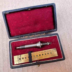 May include: A vintage metal tool with a pointed tip and a threaded end, accompanied by a rectangular brass gauge plate featuring numbered holes, both housed in a black hinged case lined with deep red velvet fabric for secure storage.