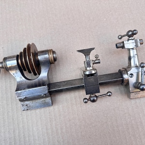 Puede incluir: Un accesorio de torno de metal que presenta un sistema de poleas escalonadas de latón a la izquierda, un soporte de herramienta central con mango ajustable y un conjunto de contrapunto a la derecha, montado sobre una barra de acero cuadrada.
