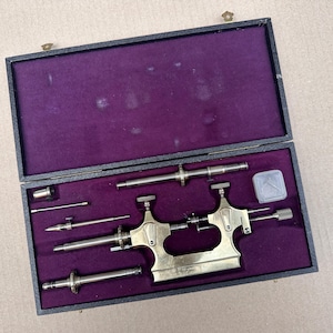 May include: A black box with a purple velvet lining contains antique watch repair tools. The tools are made of brass and steel, including a central tool and several smaller components. The box is open.