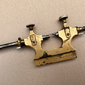 Puede incluir: Un torno de relojería de latón con un husillo de color plateado y una perilla negra. El torno está etiquetado como "STERN ANTAL & S. PHILADELPHIA, PA."