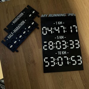 Op de afbeelding: Een zwart kunststof displaybord met witte digitale cijfers die looptijden voor 1 KM, 5 KM en 10 KM weergeven, gelabeld MY RUNNING PRS. Twee kleinere zwarte modulaire stukken zijn inbegrepen, gelabeld MARATHON en HALF-MARATHON.