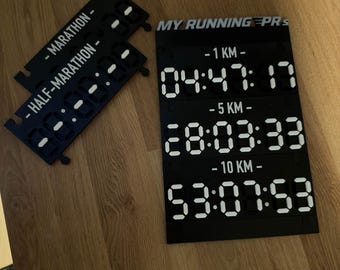 Peut inclure: Un panneau d'affichage noir en plastique avec des chiffres numériques blancs indiquant les temps de course pour 1 KM, 5 KM et 10 KM, étiqueté MY RUNNING PRS. Deux pièces modulaires noires plus petites sont incluses, étiquetées MARATHON et HALF-MARATHON.