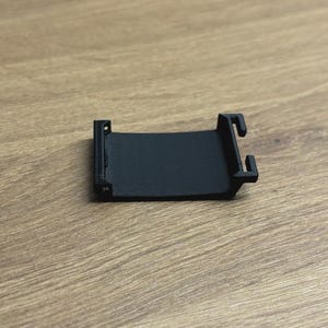 May include: A small, black, 3D-printed plastic component with a curved rectangular shape. It features two small tabs on one end and two hook-like protrusions on the other. The object is designed for a specific purpose.