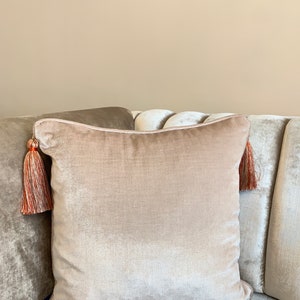 May include: A light beige velvet pillow with a tassel on each corner. The tassels are a mix of orange and beige.