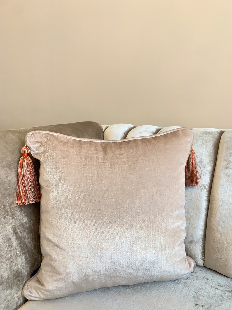 May include: A light beige velvet pillow with a tassel on each corner. The tassels are a mix of orange and beige.