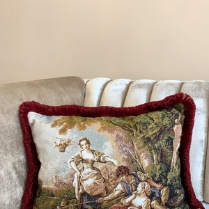 Op de afbeelding: Een decoratief kussen met een tapijtontwerp met een vrouw en twee kinderen in een tuinsetting. Het kussen heeft een bordeauxrode franjebord.