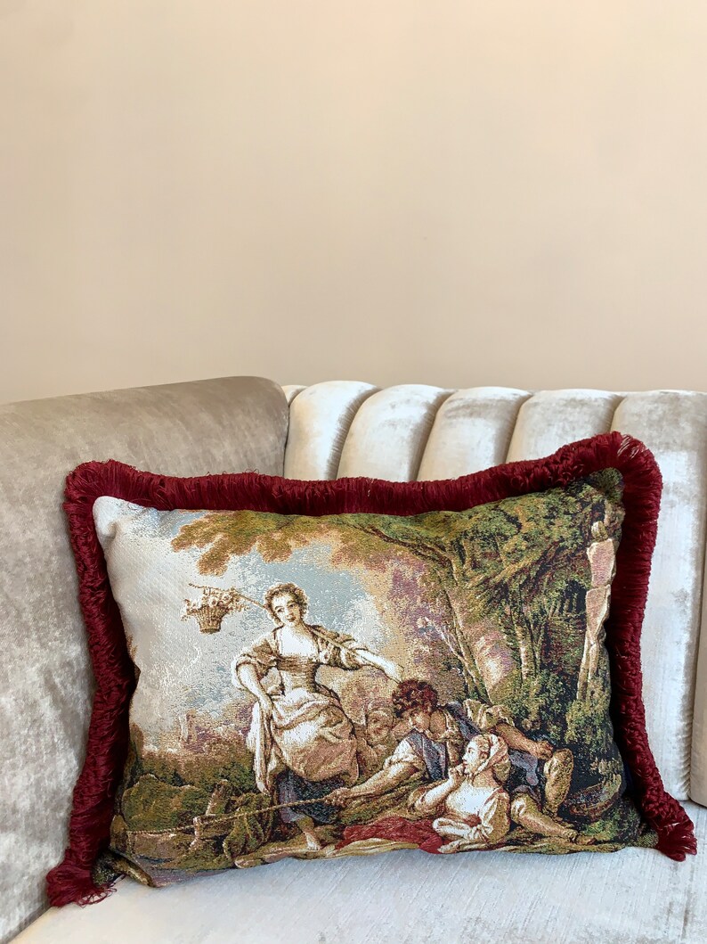 Op de afbeelding: Een decoratief kussen met een tapijtontwerp met een vrouw en twee kinderen in een tuinsetting. Het kussen heeft een bordeauxrode franjebord.