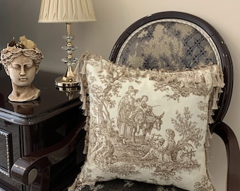 May include: A cream-colored throw pillow with a brown toile pattern featuring a scene of people and a donkey in a forest. The pillow has a fringe trim and is sitting on a brown armchair with a patterned fabric.