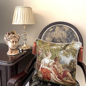 May include: A decorative throw pillow with a tapestry design featuring a couple sitting under a tree. The pillow has tassels on each corner and is resting on a chair with a patterned fabric.