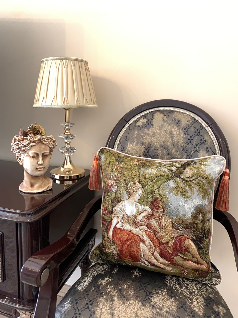 May include: A decorative throw pillow with a tapestry design featuring a couple sitting under a tree. The pillow has tassels on each corner and is resting on a chair with a patterned fabric.