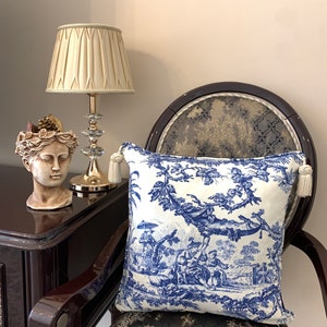 Op de afbeelding: Een blauw en wit gepatroneerd kussen met een toile-ontwerp met een pastorale scène. Het kussen ligt op een stoel met een donkerbruin houten frame en een gepatroneerde stoffen zitting.