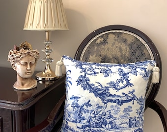 May include: A blue and white patterned throw pillow with a toile design featuring a pastoral scene. The pillow is sitting on a chair with a dark brown wooden frame and a patterned fabric seat.