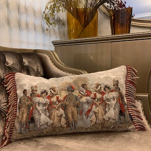 Op de afbeelding: Een decoratief kussen met een tapijtontwerp met een groep mensen in 19e-eeuwse kleding. Het kussen heeft een rode en gouden franje rand.