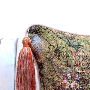 May include: A decorative throw pillow with a tapestry-like fabric featuring a green, brown, and red floral pattern. The pillow has a beige border and a long orange and white tassel.