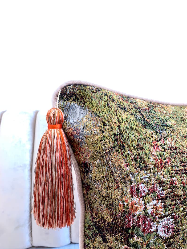 May include: A decorative throw pillow with a tapestry-like fabric featuring a green, brown, and red floral pattern. The pillow has a beige border and a long orange and white tassel.