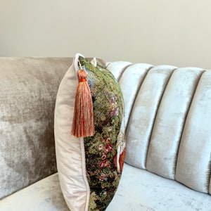 May include: A decorative throw pillow with a tapestry-like design featuring a floral pattern in shades of green, brown, and red. The pillow has a white velvet border and a long, orange tassel.